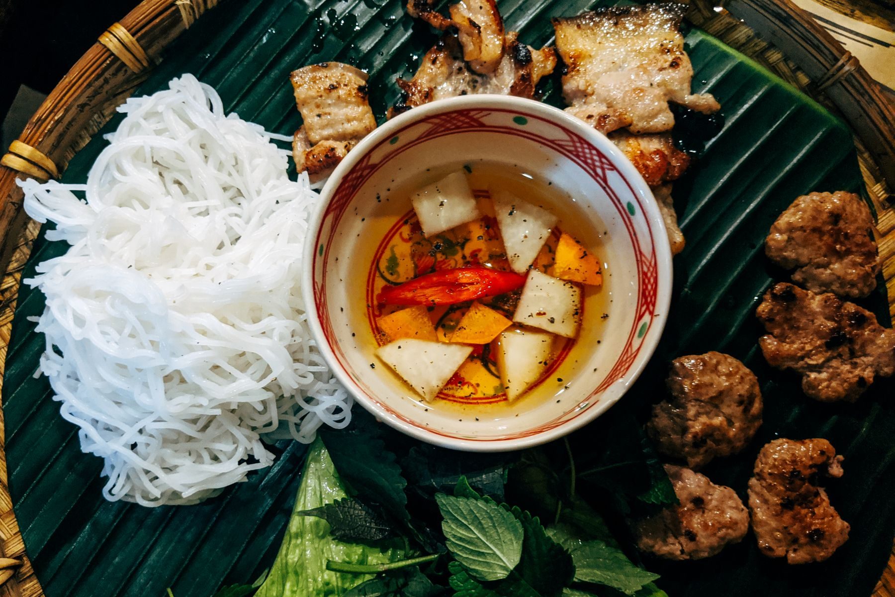 Bún chả on banana leaf: white vermicelli, charred pork patties, herbs, and the warm bowl of sauce