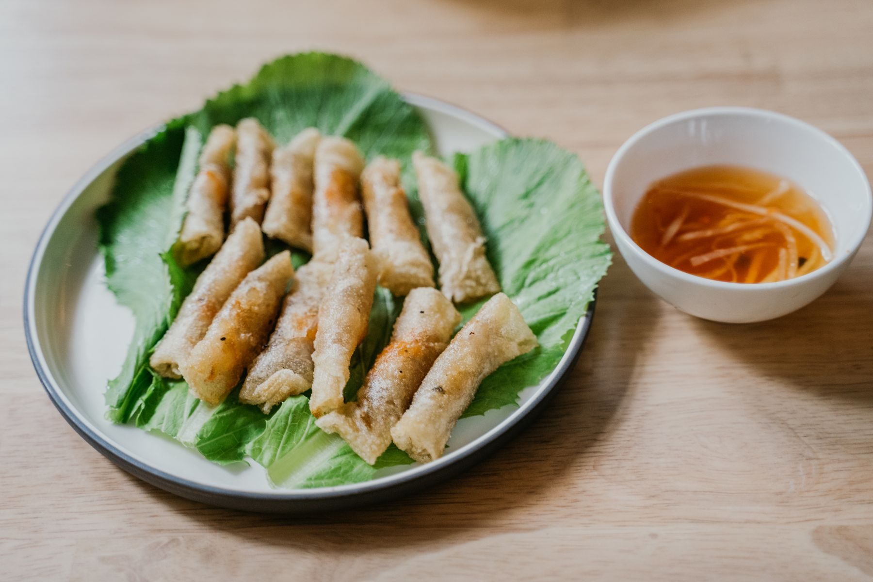 A bowl of nước chấm — amber, flecked with shredded carrot — beside a plate of nem rán